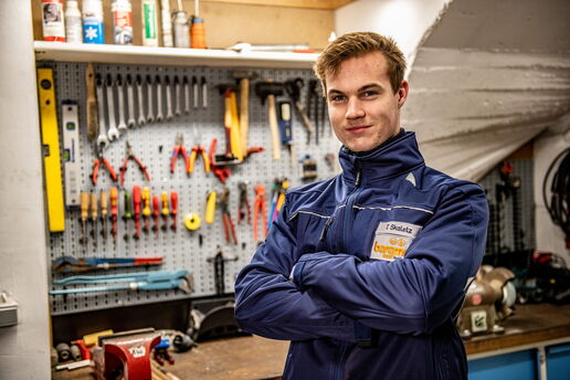 Junger Mann in blauer Arbeitskleidung steht lächelnd in einer Werkstatt vor einer Werkzeugwand mit Schraubenschlüsseln, Zangen und Werkzeugen.