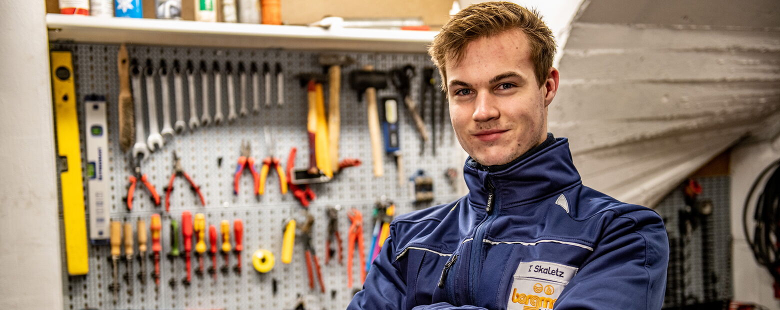 Junger Mann in blauer Arbeitskleidung steht lächelnd in einer Werkstatt vor einer Werkzeugwand mit Schraubenschlüsseln, Zangen und Werkzeugen.