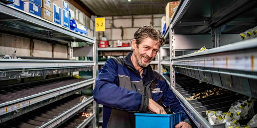 Mitarbeiter in blauer Arbeitskleidung greift lachend in eine Lagerkiste zwischen Regalen mit Kleinteilen und Verpackungen.