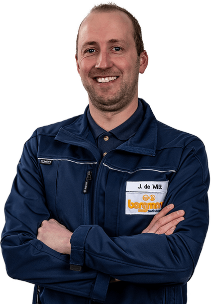 Mitarbeiter in blauer Arbeitskleidung mit Namensschild 'J. de Witt' steht mit verschränkten Armen und lächelt in die Kamera.