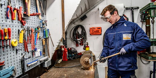 Auszubildender in blauer Arbeitskleidung bearbeitet Metall an einer Schleifmaschine in einer Werkstatt, umgeben von Werkzeugen an der Wand.