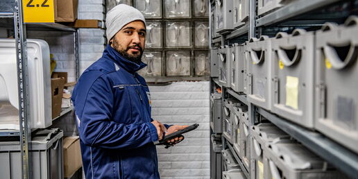 Mitarbeiter in blauer Arbeitskleidung und Mütze kontrolliert mit einem Tablet Lagerbehälter in einem Regallager.
